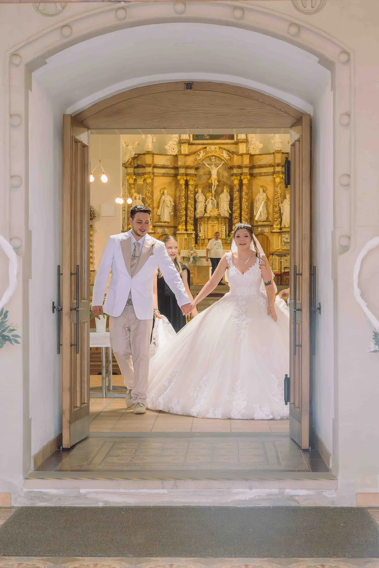 Hochzeitsreportage, das Brautpaar kommt Hand in Hand nach der Trauung aus der Kirche, von aussen durch die Eingangstür fotografiert