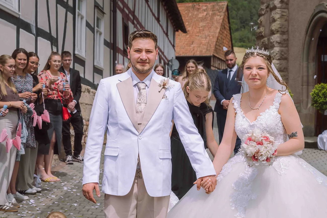 Das Brautpaar kommt Hand in Hand aus der Kirche, die Hochzeitsgesellschaft im Hintergrund