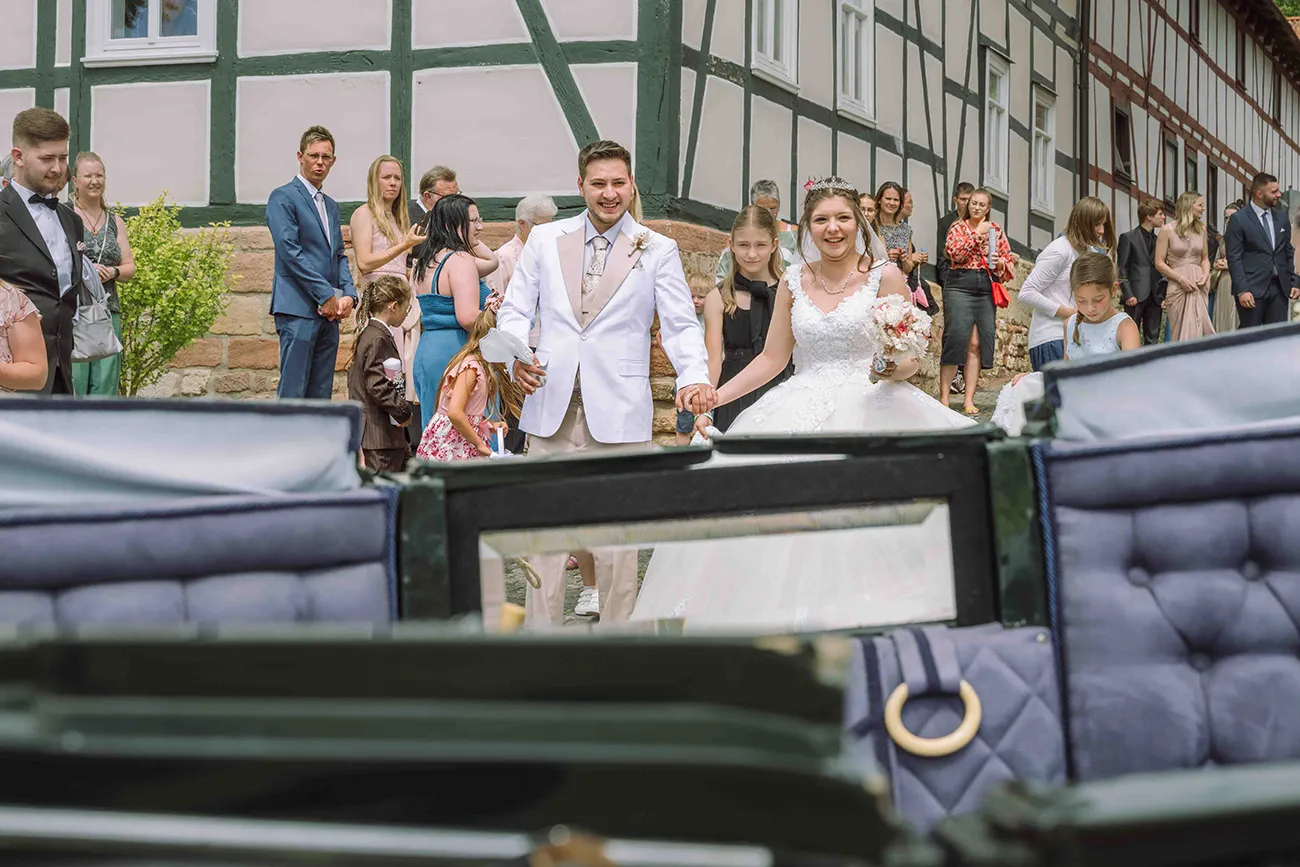 Das Hochzeitspaar geht zur Hochzeitskutsche, mit Hochzeitsgesellschaft im Hintergrund