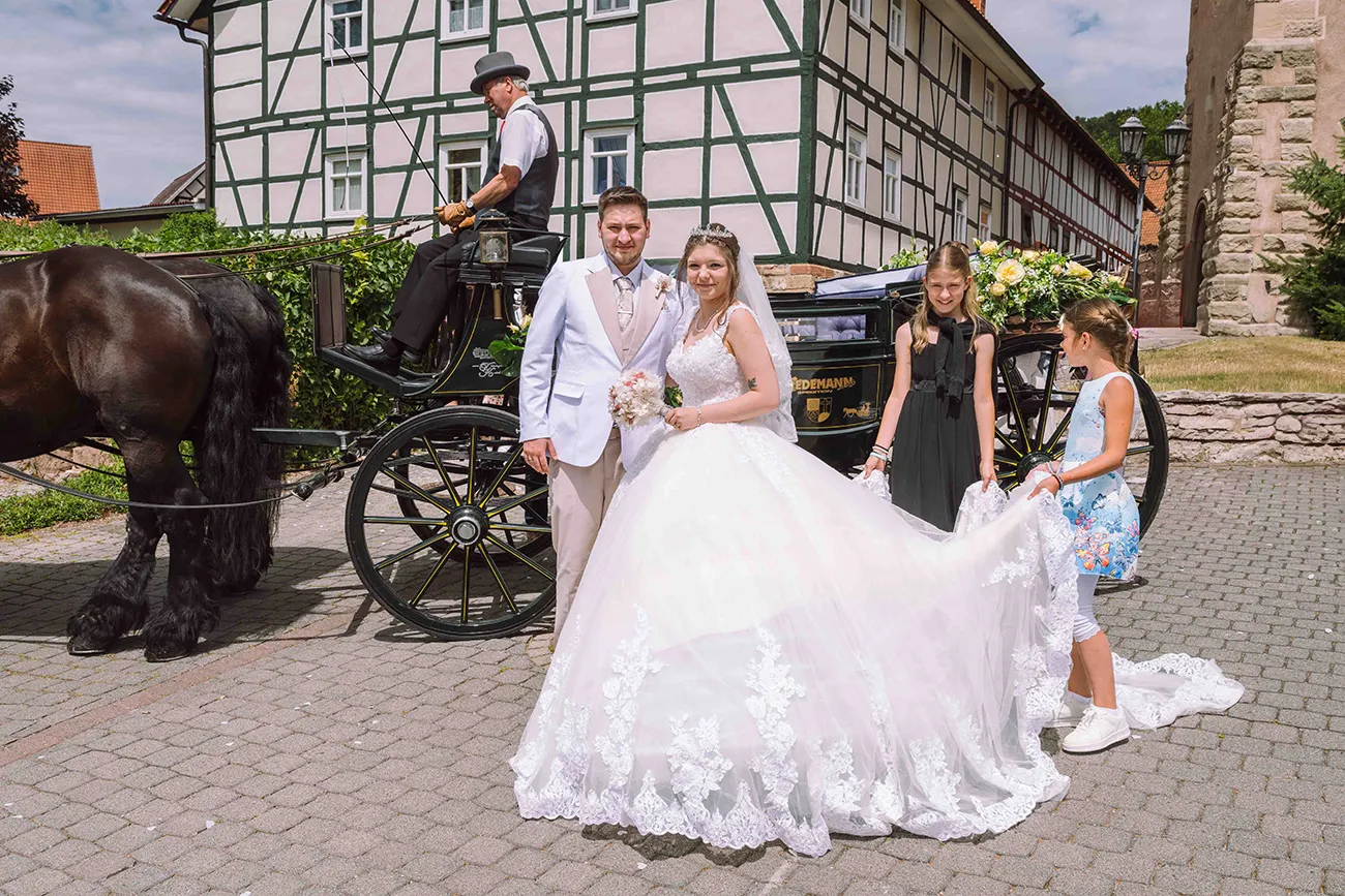 Das Brautpaar steht vor der Hochzeitskutsche im Brautkleid und weissen Anzug
