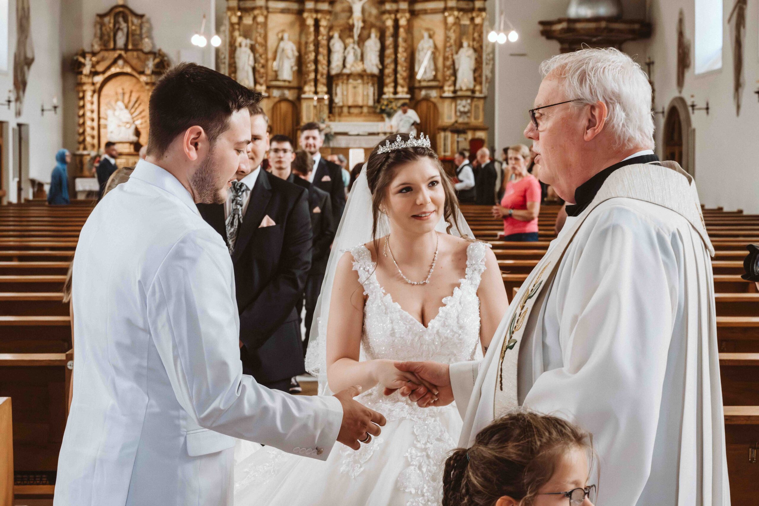 Der Pastor gratuliert als erster dem Brautpaar nach der Trauung in der Kirche. Die Hochzeitsgesellschaft im Hintergrund
