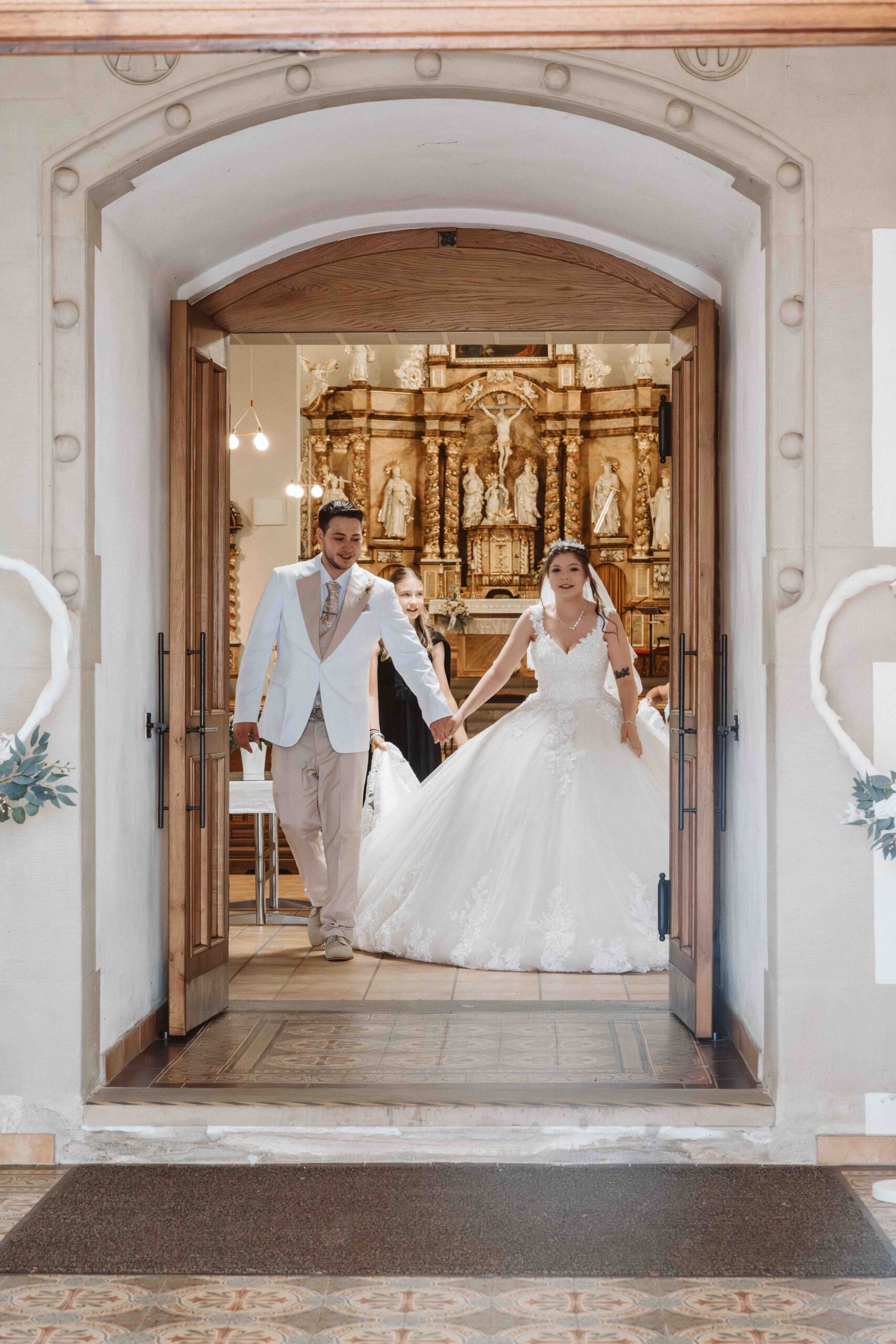 Hochzeitsreportage, das Brautpaar kommt Hand in Hand nach der Trauung aus der Kirche, von aussen durch die Eingangstür fotografiert