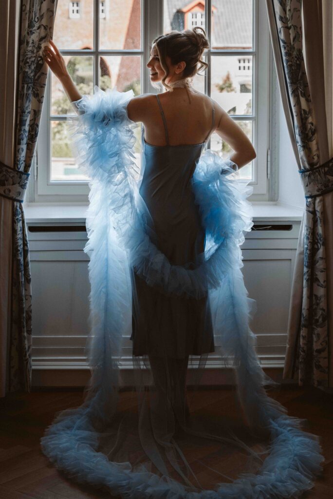 Rückenansicht einer Braut vor einem alten Fenster mit Gardinen während eines Bridal Boudoir Shootings auf Schloss Burg Heimerzheim.