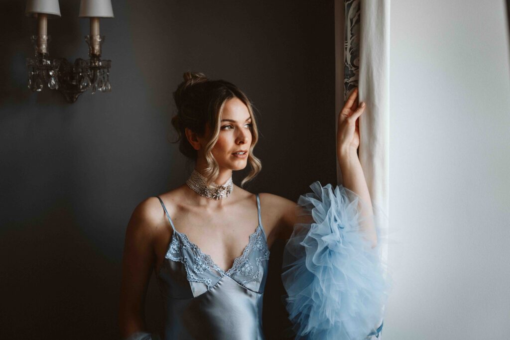 Braut in einem blauen Morgenmantel mit Federbesatz blickt während eines Editorial Styled Shootings aus dem Fenster.
