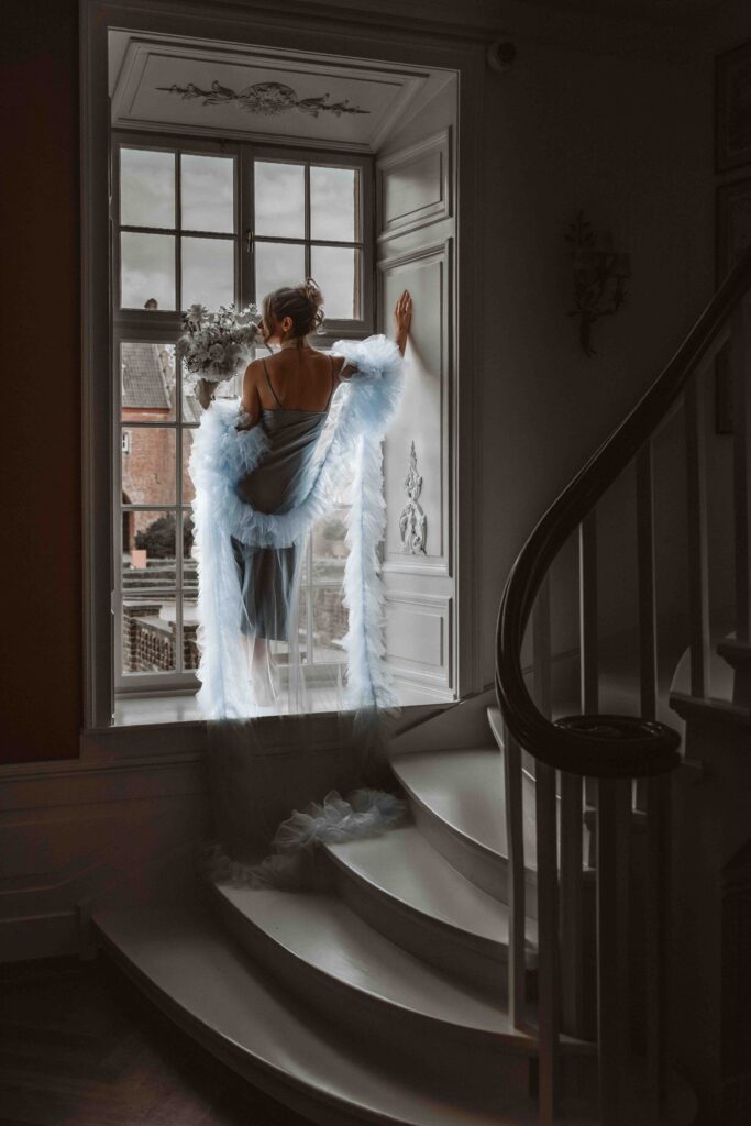 Braut im blauen Morgenmantel vor einem Fenster während eines Editorial Styled Shootings im Schloss.
