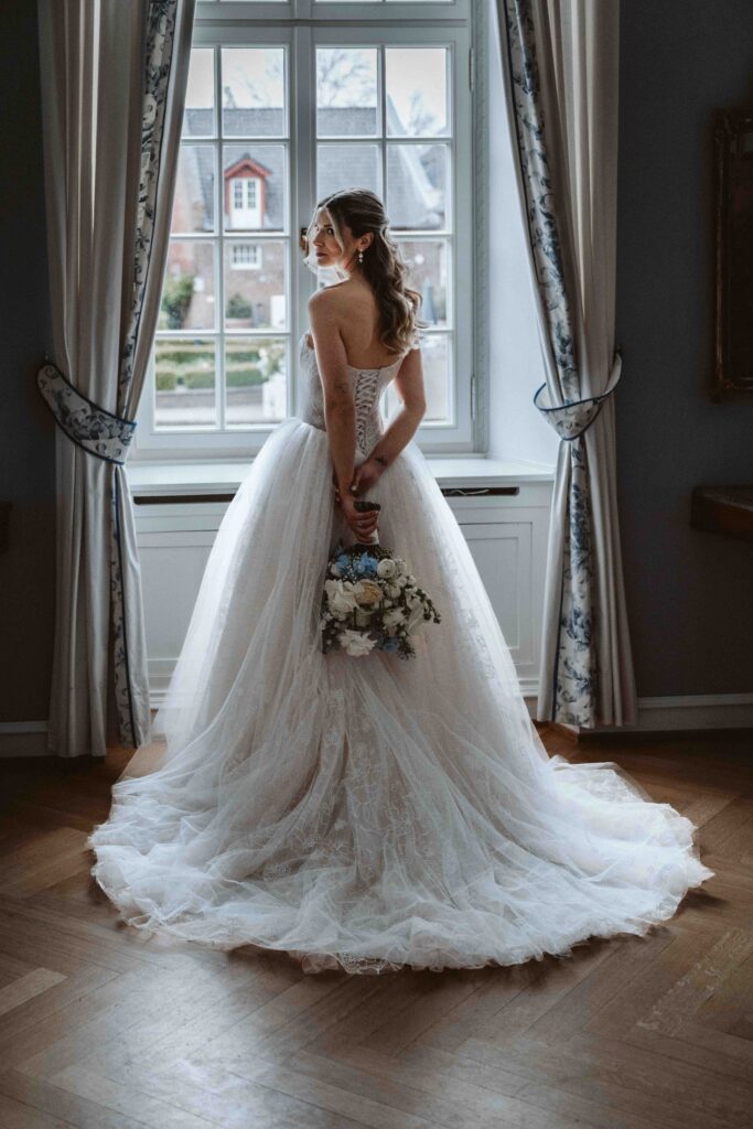 Braut mit Brautstrauß vor einem historischen Fenster auf Burg Heimerzheim kurz vor der freien Trauung. Das elegante Brautkleid mit langer Schleppe steht im Mittelpunkt der Aufnahme.