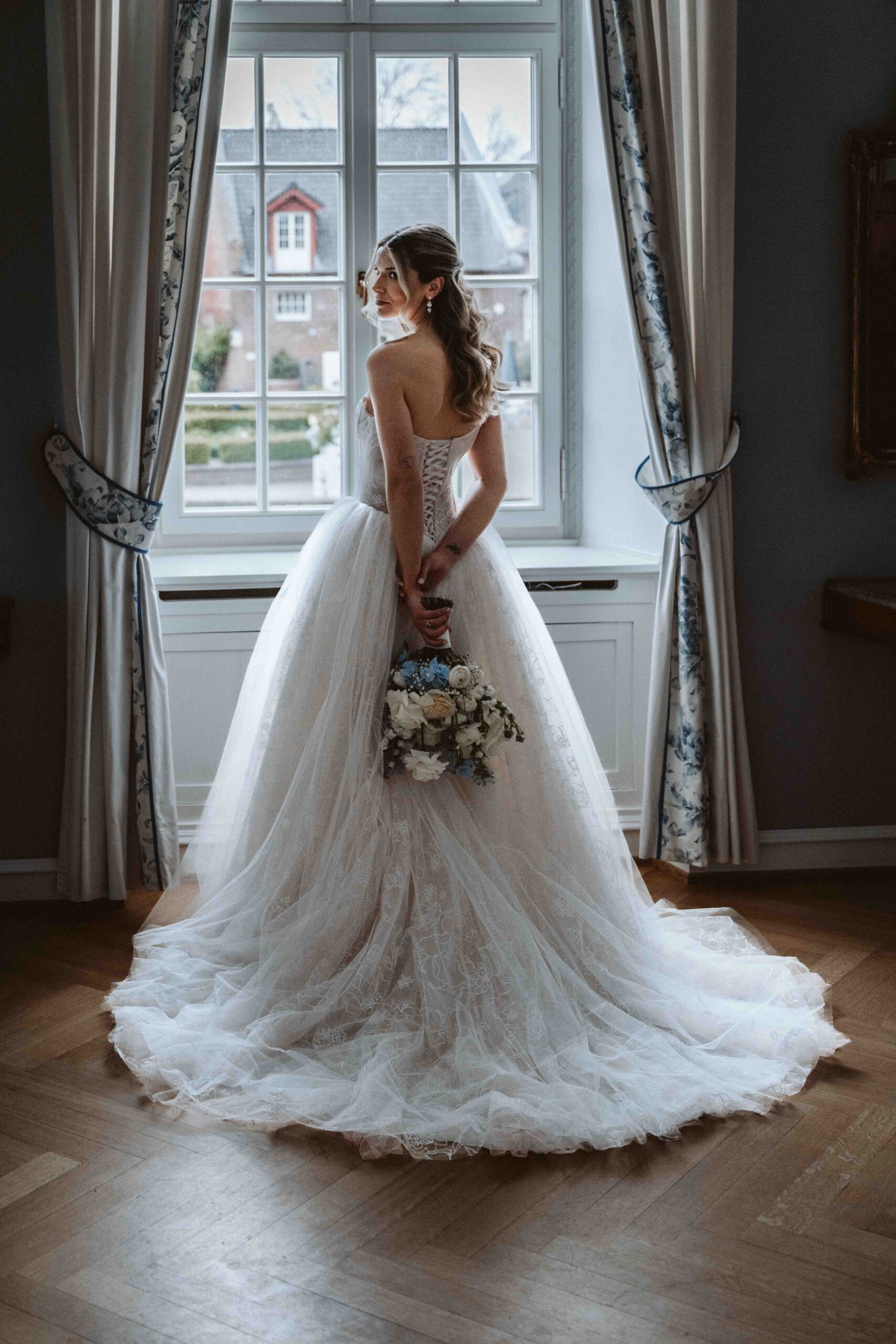 Braut mit Brautstrauß vor einem historischen Fenster auf Burg Heimerzheim kurz vor der freien Trauung. Das elegante Brautkleid mit langer Schleppe steht im Mittelpunkt der Aufnahme.
