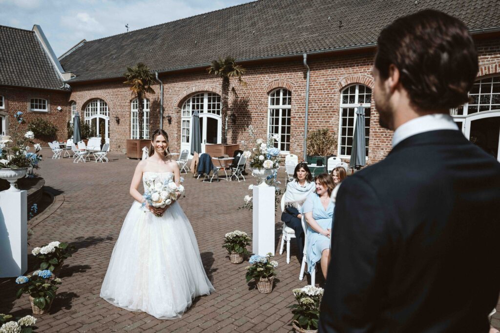 Braut schreitet während einer freien Trauung auf Burg Heimerzheim auf den wartenden Bräutigam zu, während die Hochzeitsgäste den besonderen Moment verfolgen.
