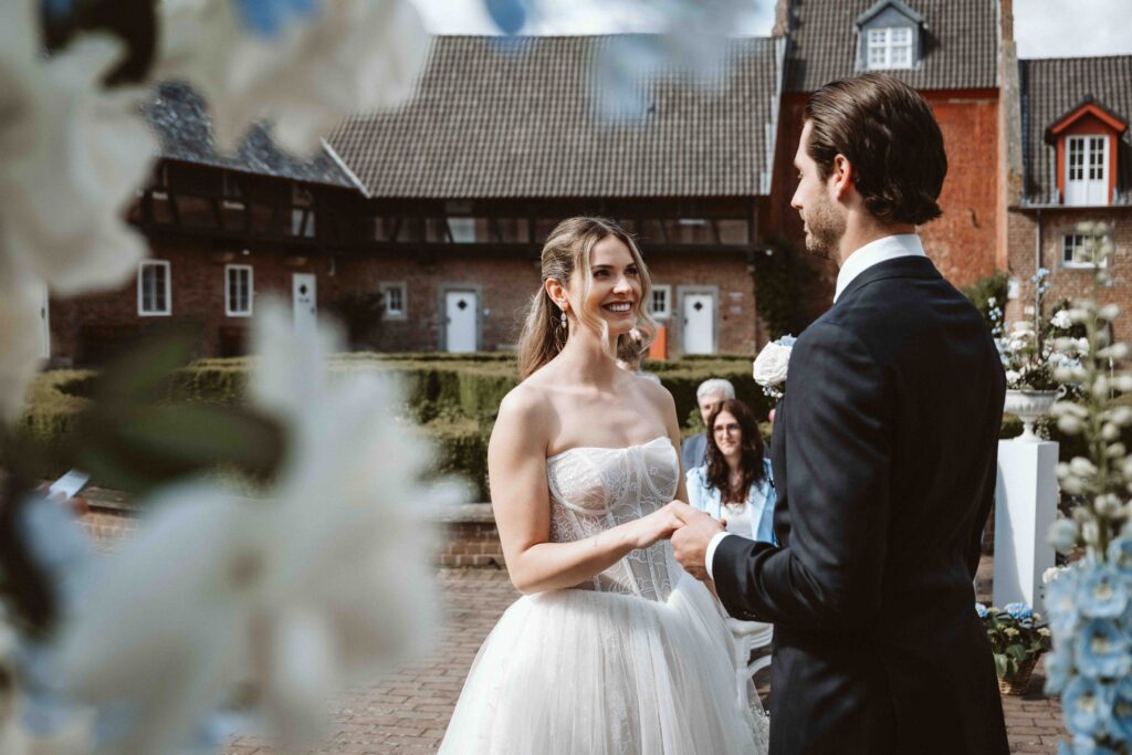 Braut und Bräutigam stehen während ihrer Hochzeitszeremonie auf Burg Heimerzheim vor dem Traubogen und schauen sich voller Freude an.