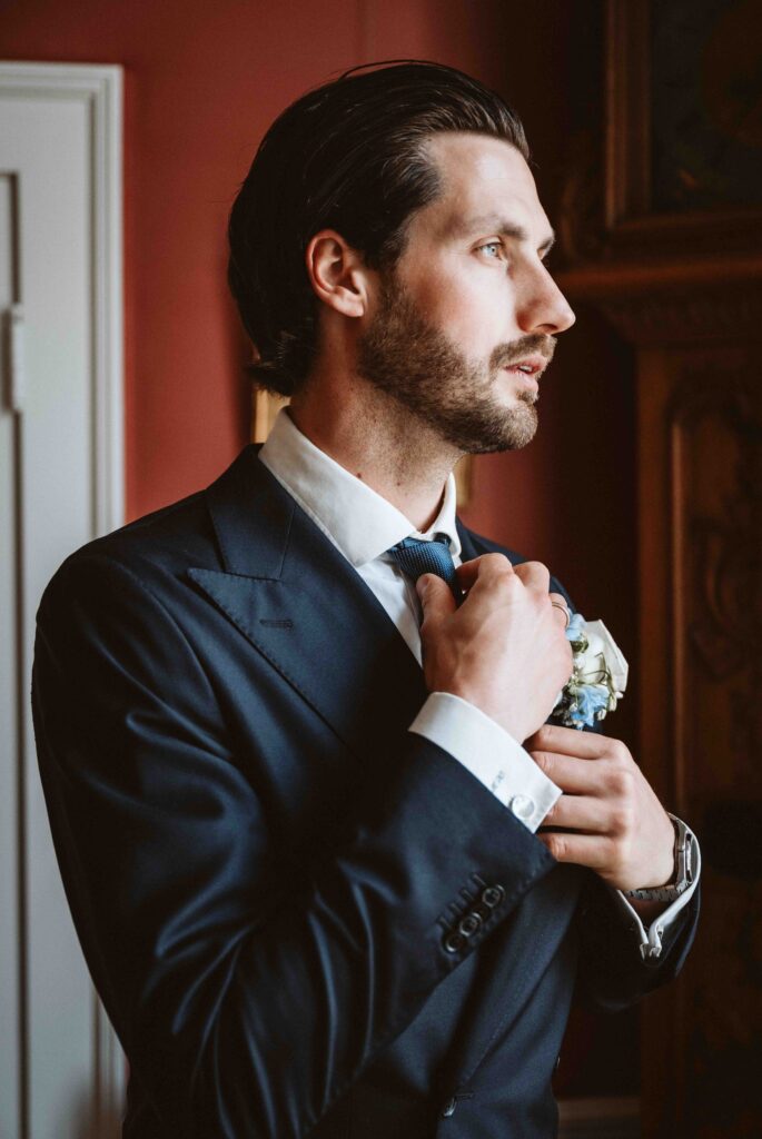 Elegantes Portrait eines Bräutigams beim Richten seiner Krawatte am Fenster im historischen Ambiente von Schloss Burg Heimerzheim.