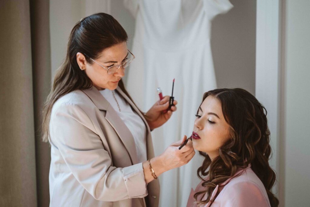 Braut beim Getting Ready mit Visagistin und Hochzeitskleid im Hintergrund
