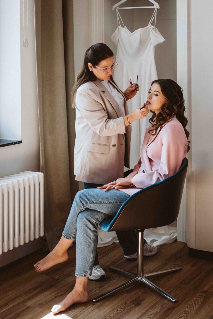 Braut beim Getting Ready in der Villa Bodinus, während die Visagistin das Make-up vollendet und das Brautkleid im Hintergrund bereit hängt.