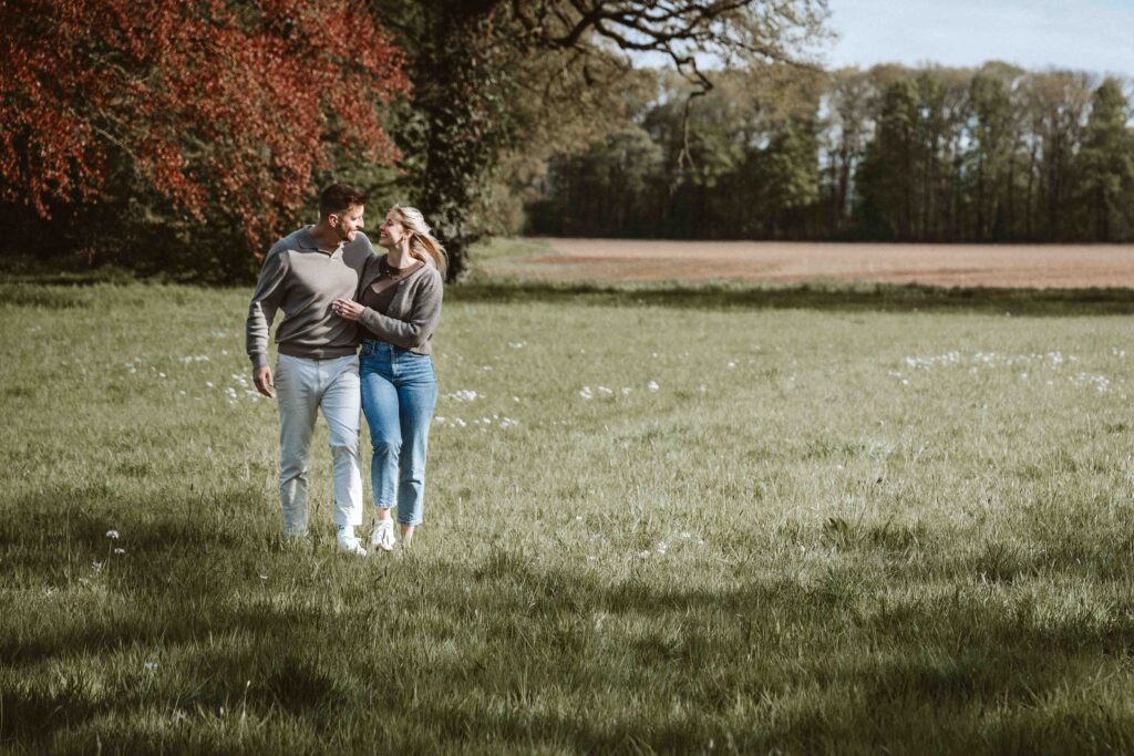 Verlobtes Paar spaziert Arm in Arm über eine weitläufige Frühlingswiese während eines natürlichen Engagement Shootings.