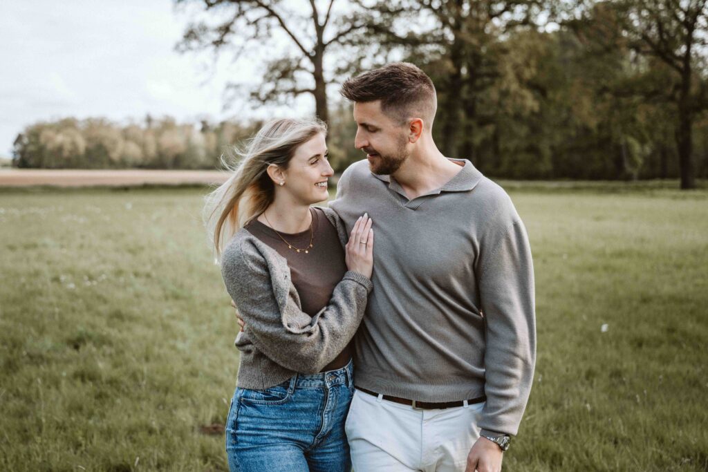 Verlobtes Paar spaziert eng umschlungen über eine Wiese und blickt sich während eines natürlichen Engagement Shootings lächelnd an.