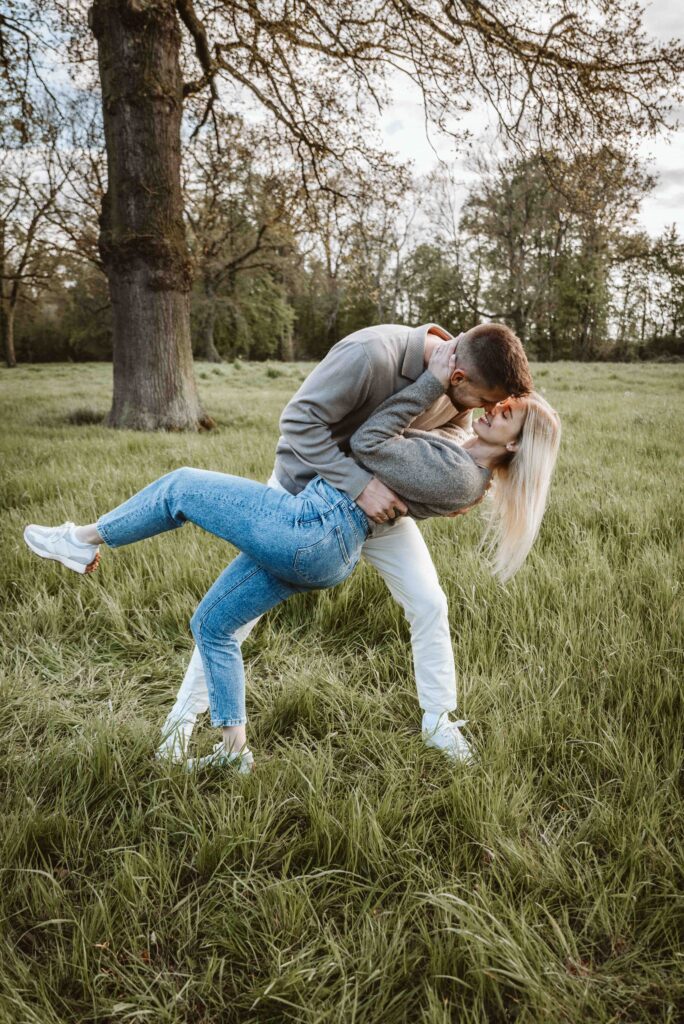 Verlobtes Paar lacht während eines verspielten Engagement Shootings auf einer Wiese im Abendlicht.
