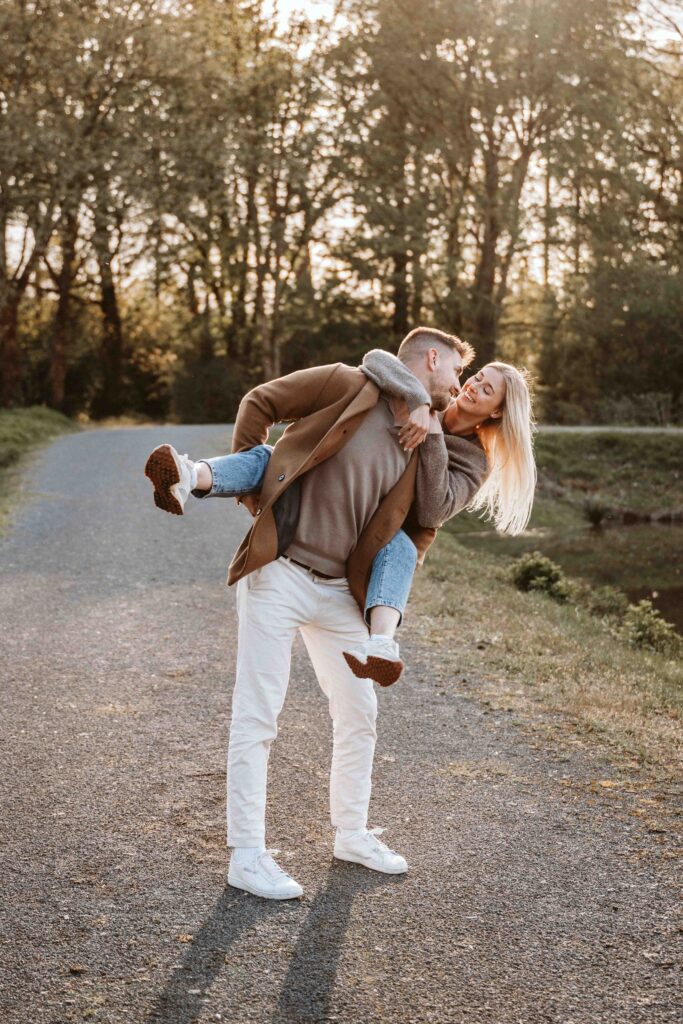 Bräutigam trägt seine Partnerin lachend während eines natürlichen Engagement Shootings im warmen Abendlicht.