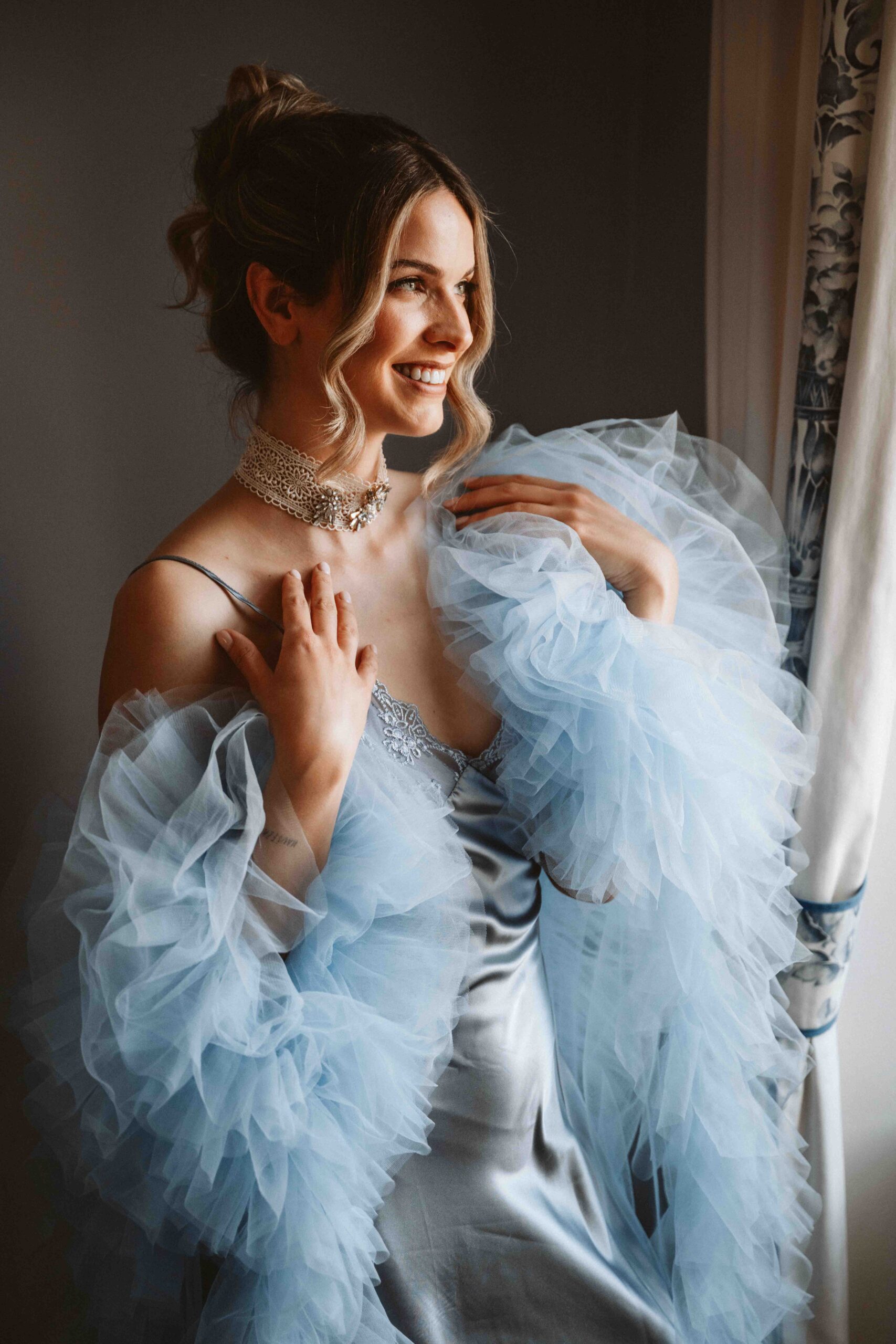 Braut lächelt während eines Boudoir-Shootings am Fenster und trägt ein elegantes Seidenkleid mit zartem Tüllmorgenmantel.