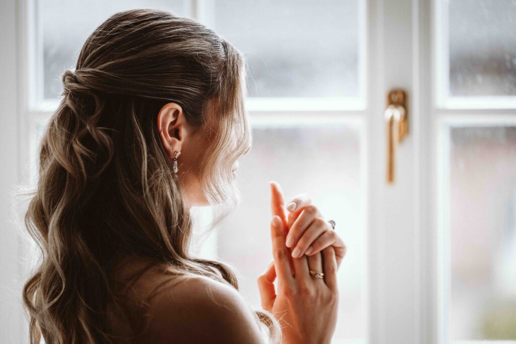Braut blickt während des Gettings Ready nachdenklich aus dem Fenster und hält ihre Hände mit Verlobungsring vor sich.