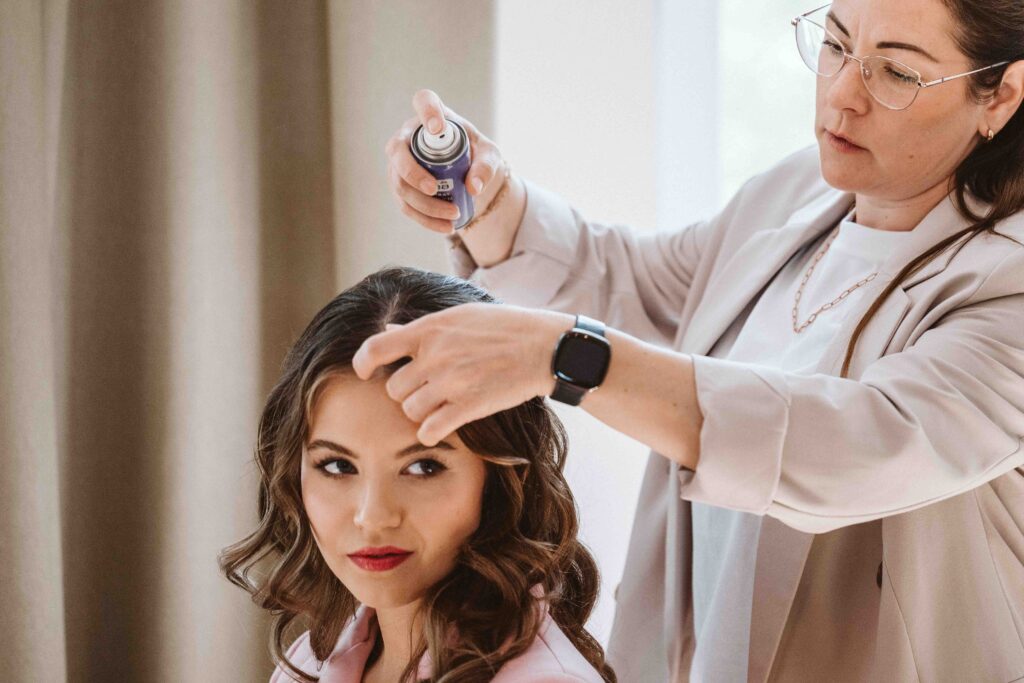 Stylistin fixiert die Frisur der Braut mit Haarspray während des Gettings Ready vor der Hochzeit.