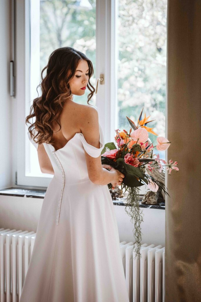 Braut im eleganten Hochzeitskleid mit tropischem Brautstrauß am Fenster der Villa Bodinus im Kölner Zoo. Die Rückenansicht zeigt das schlichte Brautkleid und die offene Schulterpartie.