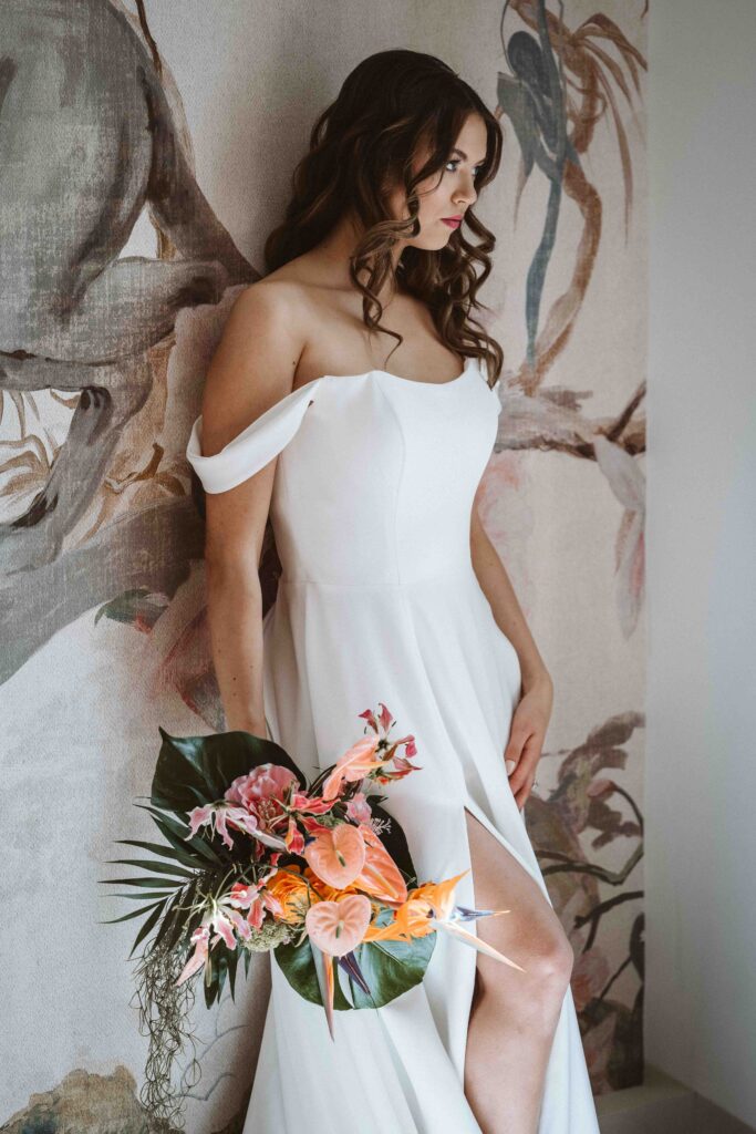 Braut im Brautkleid mit farbenfrohem Brautstrauß während des Gettings Ready in einem lichtdurchfluteten Raum.