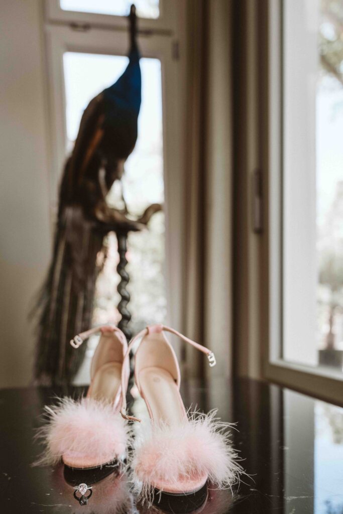 Rosafarbene Brautschuhe mit Federbesatz und Verlobungsring vor einem Pfau in der Villa Bodinus.
