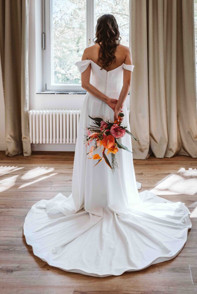 Braut in modernem Hochzeitskleid mit langer Schleppe und farbenfrohem Brautstrauß vor einem Fenster in der Villa Bodinus im Kölner Zoo.