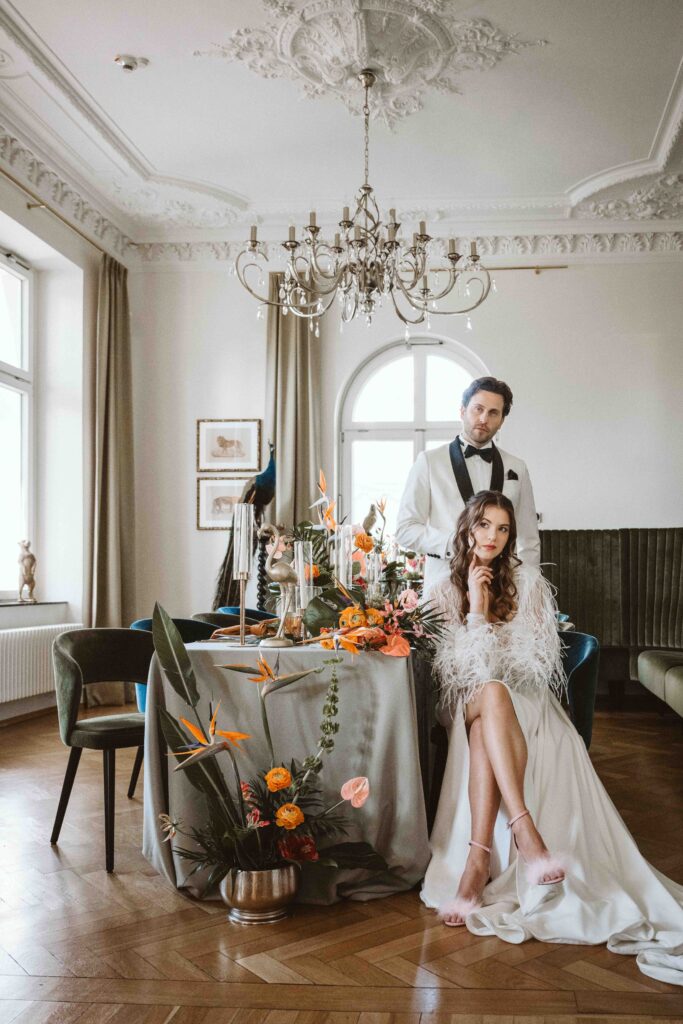 Braut sitzt an der festlich gedeckten Hochzeitstafel, während der Bräutigam hinter ihr steht – Paarshooting im eleganten Festsaal der Villa Bodinus.